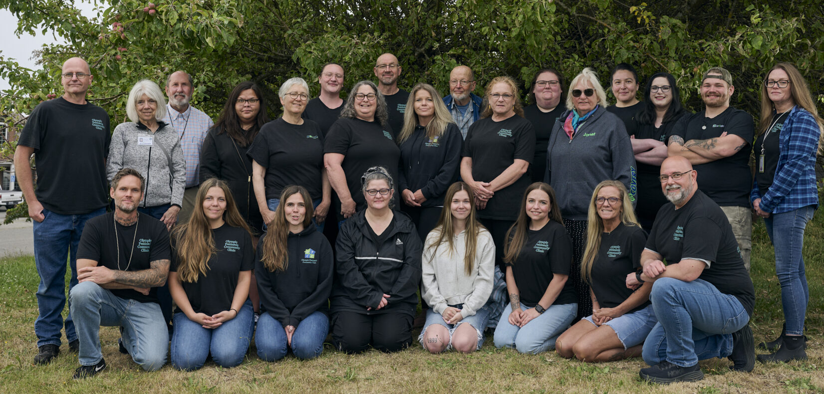 The Olympic Peninsula Community Clinic team, standing and kneeling together outdoors in front of a large tree.